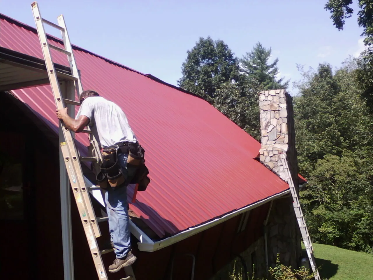 Roofer on a ladder beside a red metal roof and stone chimney — Soffit & Fascia Repair by Roofing Brothers in Ludlow